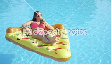 Slow Motion Close: Mutlu esmer kız ananas içeceğini yudumluyor ve şişme pizza şamandırasında dinleniyor. Bikinili genç bir kadın yaz tatilinin tadını çıkarıyor. Havuz başında kokteyl içiyor.