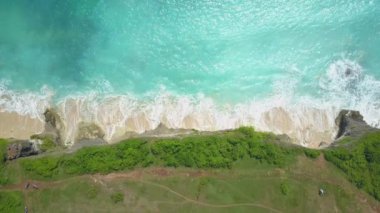 Hava üst aşağı, ağır çekim: hafifçe cliffy shore güneşli Bali Adası beyaz kumlu plaj yıkama karşı sıçramasına köpüklü whitewater okyanus dalgaları. Kayalık kaya kristal berraklığında turkuvaz denizin üzerinde yükselen