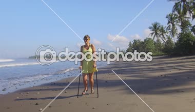 Yakın: Güçlü, tropikal yaz tatilde kum okyanus plaj boyunca koltuk değneği ile yürüyen kadın sigortalı Bali Adası. Yemyeşil palmiyeler ile deniz kıyısı boyunca strolling tatillerde yaralı turist kız