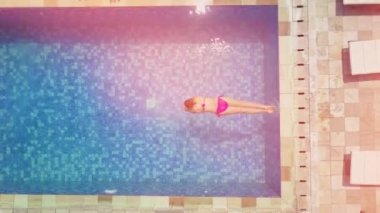 Aerial Top Down Lens: Yaz tatilinde genç bir kadın sıcak güneşli bir günde serinlemek için önce mavi havuz suyuna atlıyor. Pembe bikinili kız lüks otellerde havuz suyuna dalıyor.