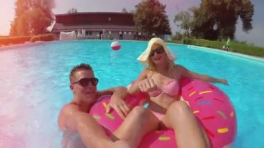Ağır çekim Lens Flare eğlenceli çift renkli floaties havuzu üzerinde arkadaşlar gülümseyen selfie video yapıyor. Mutlu gençler yaz tatile şişme pizza flamingo ve karpuz yüzen eğleniyor