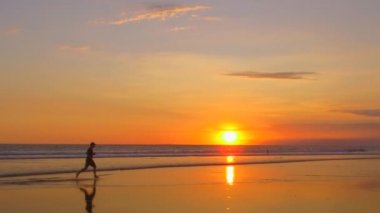 Ağır çekim: waterdrops dramatik Altın ışık günbatımında sıçramasına ıslak kumlu plajda çıplak ayakla çalışan genç adam. Dalgalanan okyanus üzerinde düz deniz kıyısı muhteşem turuncu alacakaranlıkta koşu erkek