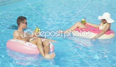 Ağır çekim: ananaslı kokteyl içme ve eğlence pembe halka floaties havuzunda rahatlatıcı genç çift. Genç adam ve kadın aşık şişme halka yüzen güneşli yaz tatil üzerinde rahatlatıcı