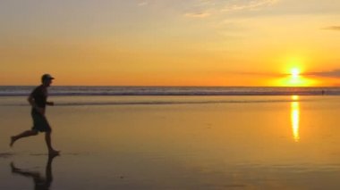 Ağır çekim: waterdrops dramatik Altın ışık günbatımında sıçramasına ıslak kumlu plajda çıplak ayakla çalışan genç adam. Dalgalanan okyanus üzerinde düz deniz kıyısı muhteşem turuncu alacakaranlıkta koşu erkek