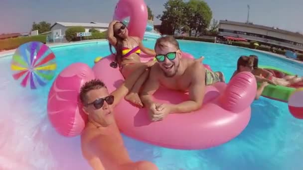 慢动作镜头耀斑有趣的家伙做自拍视频与微笑的朋友在五颜六色的浮雕在游泳池 快乐的人有水战斗在暑假充气比萨饼火烈鸟和西瓜漂浮 — 图库视频影像