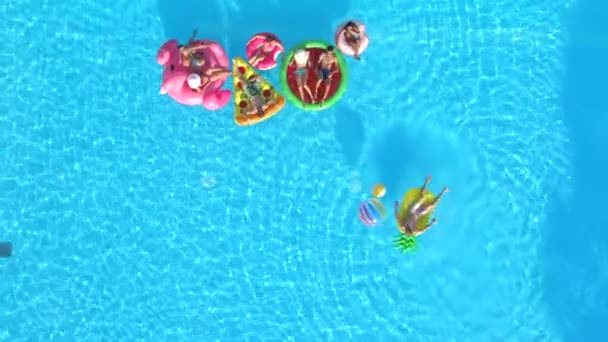空中自上而下快乐的女孩和家伙玩在五颜六色的浮雕在游泳池 微笑的朋友享受暑假在充气菠萝 比萨饼 火烈鸟 甜甜圈漂浮 — 图库视频影像