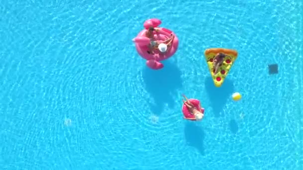 空中自上而下穿泳衣的女孩和男孩躺在有趣的充气比萨饼 甜甜圈和火烈鸟漂浮在水面上 朋友们在游泳池里享用粉色火烈鸟 甜甜圈和比萨饼 — 图库视频影像