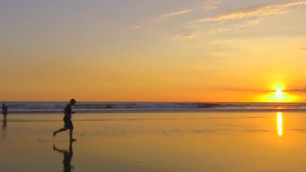慢动作 年轻人赤脚沿着光滑的沙滩跑步 在戏剧性的金色日落时 海浪飞溅着水滴 黄昏时穿着夏天衣服在海边慢跑的家伙 — 图库视频影像