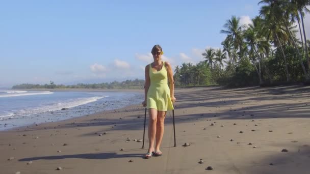 クローズ アップ 強い保険女性熱帯で夏休みに砂海のビーチに沿って松葉杖で歩くバリ島します 緑豊かなヤシの木が海岸に沿って散歩祝日無効の女の子 — ストック動画