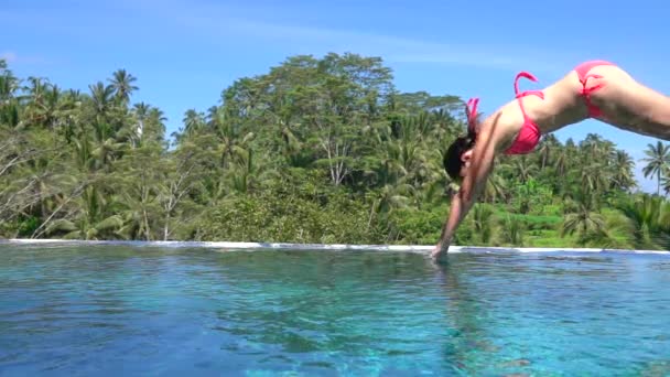 インフィニティ プールで水滴をはねかける水に頭をまず飛び込むのピンクのビキニ水着でスローモーション閉じるを若い女性 日当たりの良いバリ島のパーム ツリー ジャングル リゾートで夏休みを楽しんでいる女の子 — ストック動画