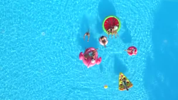 Antenne von oben nach unten: fröhliche Mädchen und Jungs, die mit Ball auf Schwimmern im Pool spielen. fröhlich lächelnde Freunde genießen den Sommerurlaub auf aufblasbaren Ananas-, Pizza-, Flamingo-, Wassermelonen- und Donut-Wagen