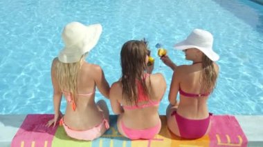Ağır çekim yakın çekim: Üç neşeli kız havuz kenarında oturan ve güneşli günde kokteyli içki içme pembe bikini. Üç genç kadın gülümseyerek ve şemsiyeli içkilerden ile kızartma yaz tatil