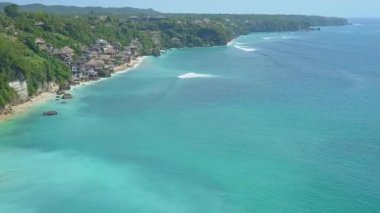 Hava Vertigo etkisi uçan güzel lüks turizm tesisi derin mavi denize bakan cliffy sahil şeridi üzerinde oceanfront kabin evleriyle yukarıda. Köpüklü deniz dalgalar güneşli Bali egzotik plaj yıkama
