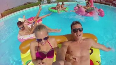 Ağır çekim Lens Flare Selfie: çift ve renkli floaties havuzu üzerinde zevk arkadaşları gülümseyerek. Flamingo, karpuz ve halka yaz su şişme pizza üzerinde mücadele, mutlu gençler yüzen