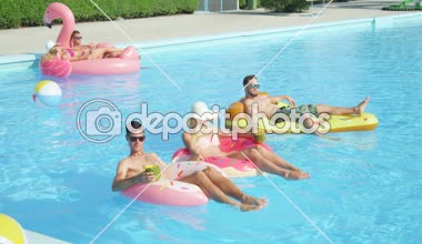 Slow Motion: Rahatlamış gençler gülümsüyor ve güneşli yaz güneşinin tadını çıkarıyorlar havuzda şişme şamandıralarda güneşleniyorlar. Neşeli arkadaşlar şişme pizza, çörek ve flamingo şamandıralarıyla yatıp içiyorlar.