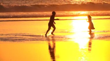 Rüya gibi kumlu Kuta Beach sunny Bali harika Altın ışık günbatımında oynayan yakın kadar ağır çekim siluet neşeli çocuklar. Güneş doğarken waterdrops sıçramasına sığ okyanus su içinde çalışan çocuk