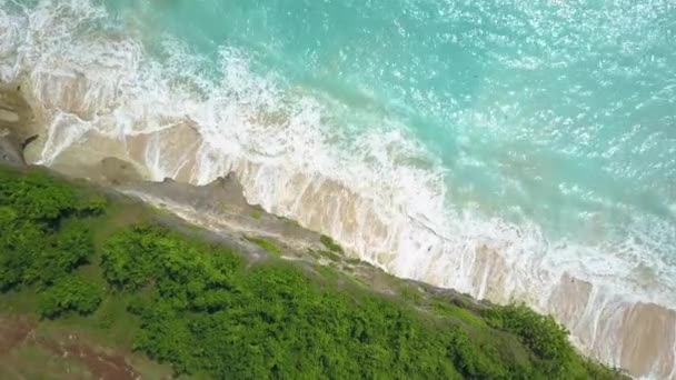 空中顶向下 泡沫白水海浪轻轻飞溅对岩壁海岸洗涤白色沙滩在阳光明媚的巴厘岛岛 岩石岩壁上升起的水晶清澈的翡翠海 — 图库视频影像