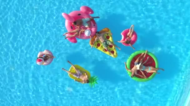 Antenne von oben nach unten: verspielte Mädchen und Jungs, die auf bunten Schwimmanlagen im Pool liegen. glücklich lächelnde Freunde genießen den Sommerurlaub auf aufblasbaren Ananas, Pizza, Flamingo, Wassermelone und Donut schwimmt
