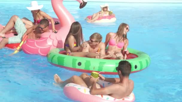 慢动作特写快乐微笑的学生有水枪战斗在五颜六色的浮雕在春假的泳池派对上 快乐的年轻人在充气火烈鸟 西瓜和甜甜圈上玩耍 — 图库视频影像