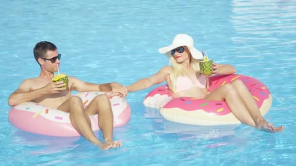 Zeitlupe: junges Paar trinkt Ananascocktails und entspannt auf lustigen rosafarbenen Donut-Floaties im Pool. Verliebte junge Männer und Frauen entspannen auf aufblasbaren Donut-Schwimmern im sonnigen Sommerurlaub