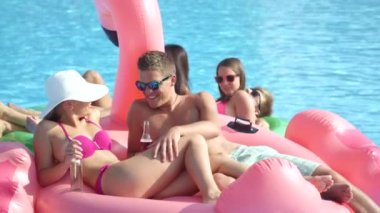 Slow Motion Close: Birbirine aşık gülümseyen, içki içen ve havuzda yüzen pembe flamingoya uzanan çift. Kız arkadaş ve erkek arkadaş, şişme flamingo şamandırasında arkadaşlarıyla güneşli bir yaz geçiriyorlar.