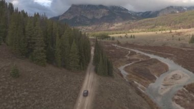 Anteni: çamurlu yol yalak üzerinde araba siyah Suv araba güzel Nehri Vadisi çam ağaçları görkemli kayalık Dağları Wyoming ABD doğru geçmiş. Off road jeep bulutlu Dağları altında çakıl yolda seyahat