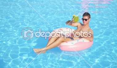 Slow Motion Close: Mutlu adam ananas içeceğini yudumluyor ve şişme çörek arabasında dinleniyor. Genç adam yaz tatilinin tadını çıkarıyor. Havuz başında kokteyl içiyor.