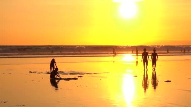 Ağır çekim yakın kadar siluet Altın ışık günbatımı kum delik kazma çocuklara yaz tatillerini Kuta beach Bali üzerinde oynama. Aileler ve çocuklar okyanusta yüzmeye. Büyük dalgalar güneş doğarken sıçramasına