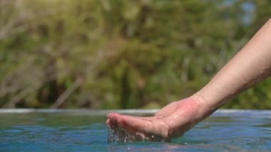 Ağır çekim yakın çekim: su ile oynarken, parmakları ile yakalamak ve orman resort sonsuzluk havuzunda içine dökülen beyaz el. İnsan palmiye su, scooping damla su yüzeyine düşen