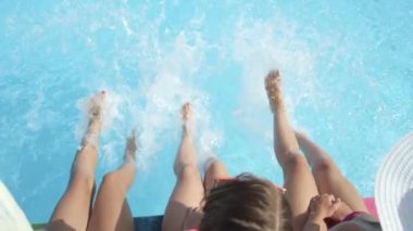 Ağır çekim yakın çekim: sıcak güneşli günde ayakları ile su sıçramasına havuzu kenarında oturan üç tanınmaz neşeli kız. Üç oynak genç kadın su bacaklar ile yaz tatili