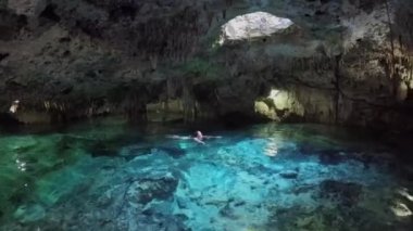 Tavandaki delikten gelen güneş ışığıyla sihirli yeraltı mağarasında turkuaz havuzda yüzen genç bir kadın. Aktun Chen 'deki sarkıtların ve dikitlerin büyüleyici mimarisi.