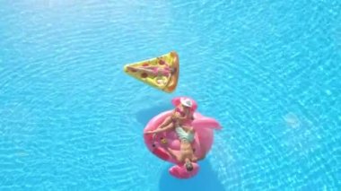 Aerial Top Down Lens Fişeği: Mayo giymiş genç bir adam ve kadın şişme pizza ve flamingo üzerinde suda yüzüyorlar. Mutlu arkadaşlar tatilde pembe flamingoyla eğleniyor ve pizza havuzda yüzüyor.