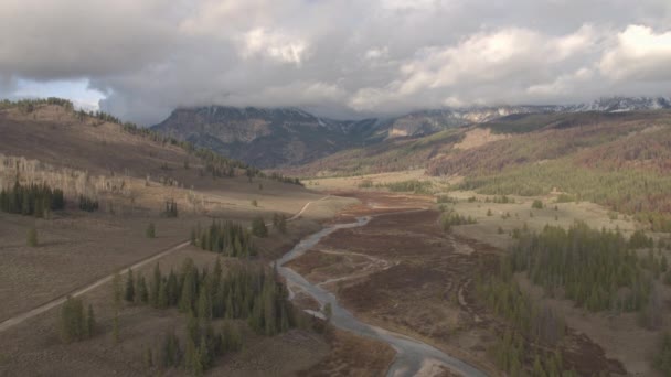 AÉRIAL : Voler au-dessus de la belle vallée fluviale au-dessus des pins vers les majestueuses montagnes Rocheuses dans le Wyoming, États-Unis. Chemin de gravier menant vers les montagnes de Grand Teton par une journée ensoleillée d'automne 