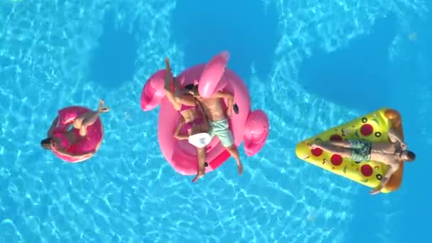 空中自上而下的微笑的女孩和男孩在泳衣铺设在有趣的充气比萨饼 甜甜圈和火烈鸟漂浮在水面上 快乐的朋友享受粉红色的火烈鸟 甜甜圈和比萨饼漂浮在游泳池 — 图库视频影像