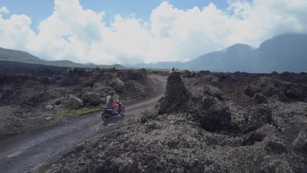空中クローズアップ壮大なキンタマニ火山 インドネシアに向かってバトゥール山の風光明媚な石の黒い溶岩の道に沿って若い男と女性がスクーターに乗っています カルデラへのロードトリップ中の観光客 — ストック動画