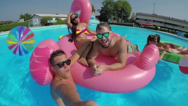 缓慢的运动自拍有趣的家伙做自拍视频与微笑的朋友在五颜六色的浮雕在游泳池 快乐的青少年有水打架在暑假充气比萨饼火烈鸟和西瓜漂浮 — 图库视频影像