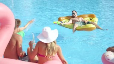Slow Motion Close Up: Neşeli gülümseme grubu havuz partisinde yaz tatilinin tadını çıkarıyor. Neşeli gençler şişme pizza, flamingo ve donut şamandıralarıyla eğleniyorlar.