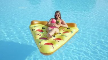 Slow Motion Close: Mutlu esmer kız ananas içeceğini yudumluyor ve şişme pizza şamandırasında dinleniyor. Bikinili genç bir kadın yaz tatilinin tadını çıkarıyor. Havuz başında kokteyl içiyor.