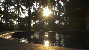 Kadar yakın, ağır çekim: lüks turizm tesisi güneşli Bali yüzme havuzunda camsı su yüzeyinde yansıtan altın güneş. Güzel bahçesi olan yemyeşil palmiye ağaçlarının arka bahçesinde muhteşem Villa