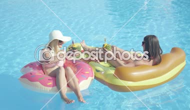 Slow Motion Close Up: İki gülümseyen kız arkadaş renkli şamandıralarda uzanıyor, yaz tatilinde içkiyle kadeh kaldırıyorlar. Kokteyllerini yudumlayan, şişme pizza ve pembe donut yiyen genç kadınlar.