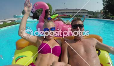 Ağır çekim Lens Flare Selfie: çift ve renkli floaties havuzu üzerinde zevk arkadaşları gülümseyerek. Flamingo, karpuz ve halka yaz su şişme pizza üzerinde mücadele, mutlu gençler yüzen