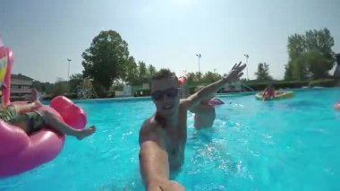Ağır çekim Selfie Lens Flare: selfie video havuzunda renkli floaties üzerinde arkadaşlarınızla yapan eğlenceli adam. Mutlu insanlar su şişme pizza flamingo ve karpuz yüzen yaz tatile sıkı mücadele var