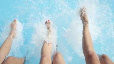 Ağır çekim yakın çekim: sıcak güneşli günde ayakları ile su sıçramasına havuzu kenarında oturan üç tanınmaz neşeli kız. Üç oynak genç kadın su bacaklar ile yaz tatili