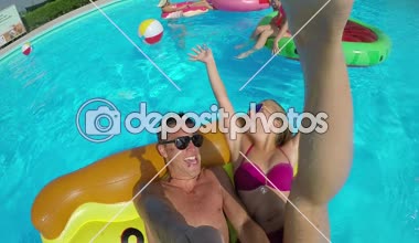 Slow Motion Selfie: Neşeli renkli şamandıralarla oynayan gülümseyen gençler. Şişme pizza, flamingo, karpuz ve donut yiyen mutlu gençler havuz partisinde su sıçratıyor.