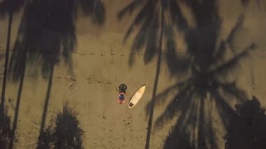 Hava yukarıdan aşağıya Distancing: Sıçramasına köpüklü okyanus su dalgaları arıyorum güzel kahverengi kumsalda oturuyor surfboard ile genç adam. Güneşli Bali güzel tropik sahil üzerinde rahatlatıcı surfboarder