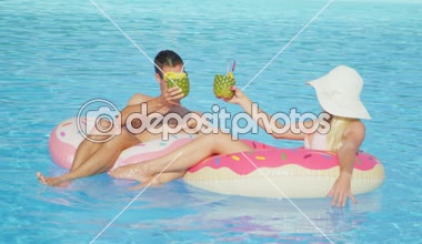 Ağır çekim: ananaslı kokteyl içme ve eğlence pembe halka floaties havuzunda rahatlatıcı genç çift. Genç adam ve kadın aşık şişme halka yüzen güneşli yaz tatil üzerinde rahatlatıcı