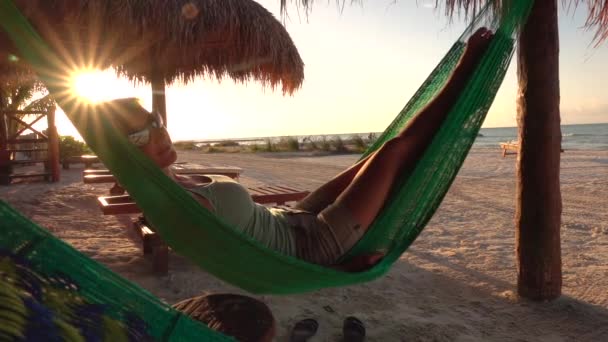 慢动作特写漂亮女孩暑假躺在吊床上摆动在美丽的沙滩在金光日出遮阳伞 戴着太阳镜在海边睡觉的迷人女人 — 图库视频影像