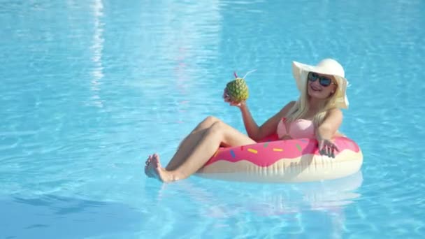 慢动作特写快乐的金发女孩喝菠萝饮料 放松充气粉红色甜甜圈浮动 穿着比基尼的年轻女子在泳池里享受暑假喝粉色的鸡尾酒 — 图库视频影像