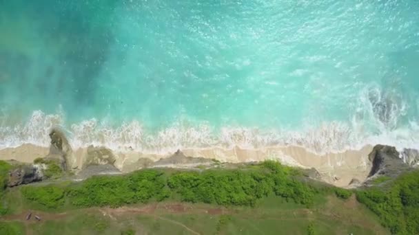空中顶向下 温和的泡沫白水海浪溅在阳光明媚的巴厘岛岩壁海岸洗涤白色沙滩 郁郁葱葱的岩石墙上升起的水晶清澈的翡翠海 — 图库视频影像