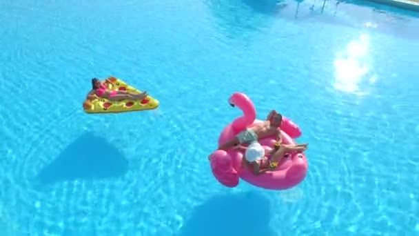 穿着泳衣的年轻女孩和男孩穿着有趣的充气比萨饼和粉色火烈鸟漂浮在水面上 假期快乐的朋友们在游泳池里享用粉色火烈鸟和比萨饼 — 图库视频影像